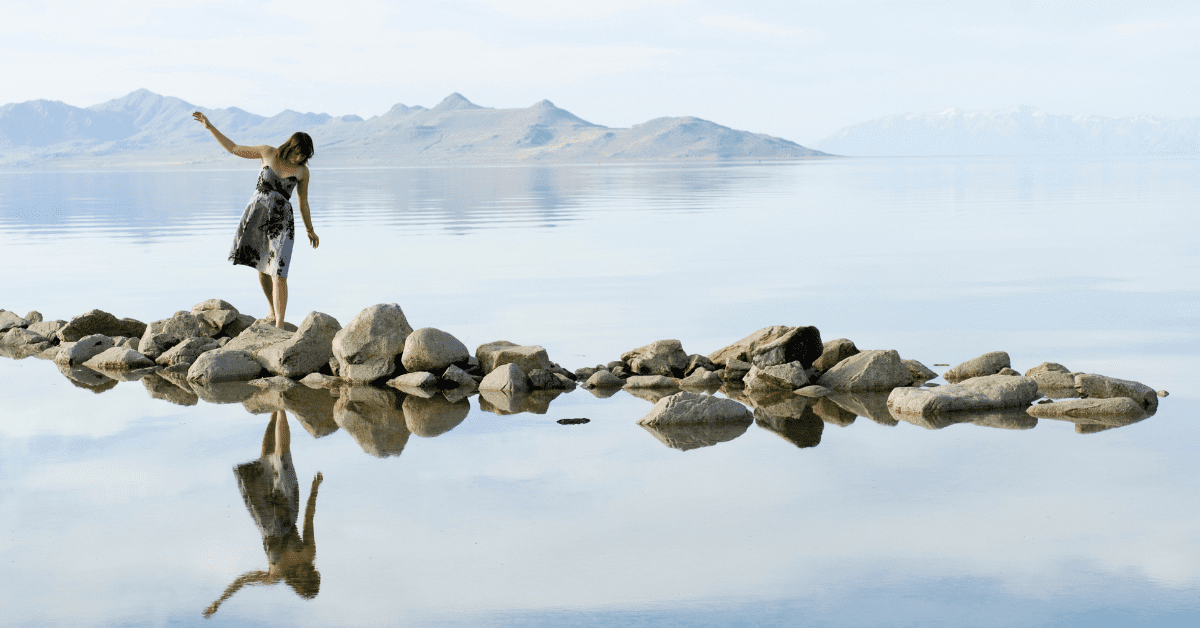 Photograph of a woman balancing on some rocks over a body of water with her reflection clearly below