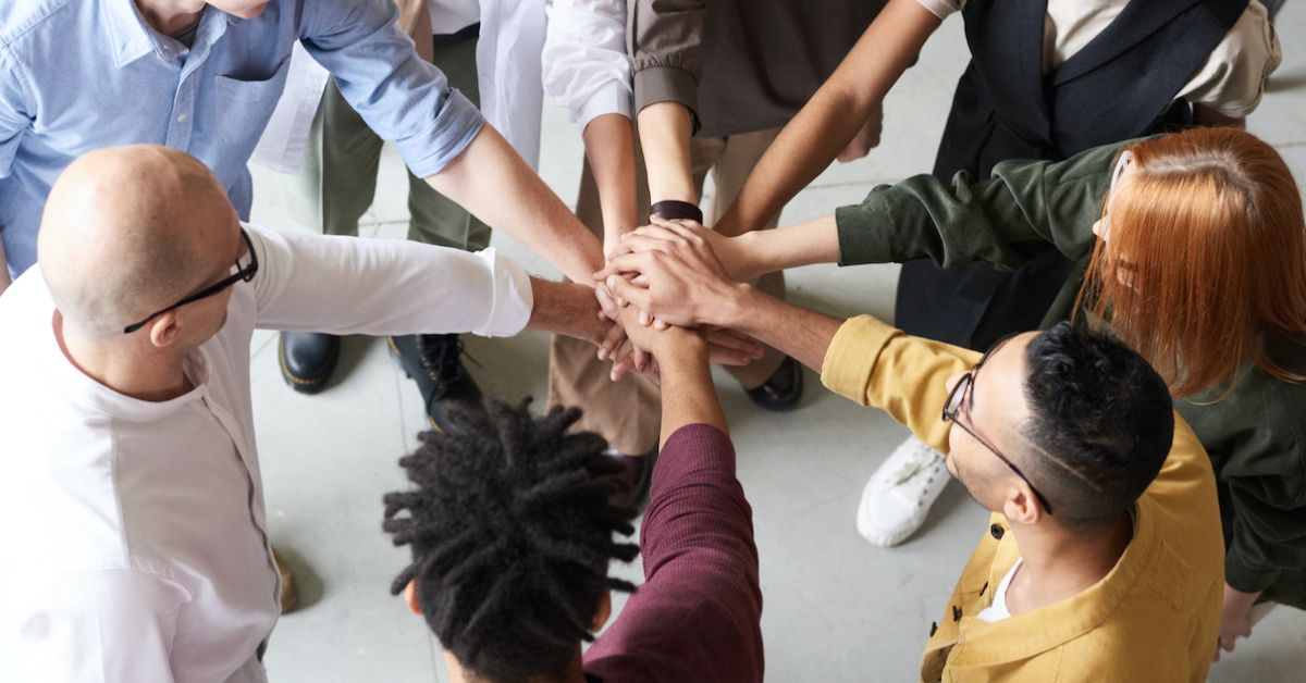 Image is of a circle of adults with their hands in the middle touching, as if doing a cheer.