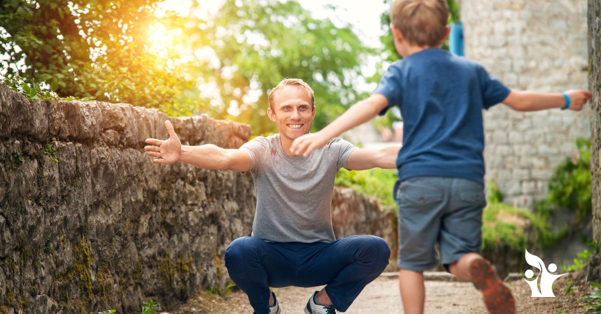 Image is of a man squatting down to give a hug to a little boy who is running towards him.