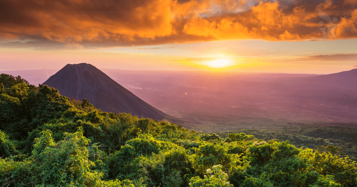 Photograph of a volcano at sunset with lush trees around its base and an angry orange cloudy sky above