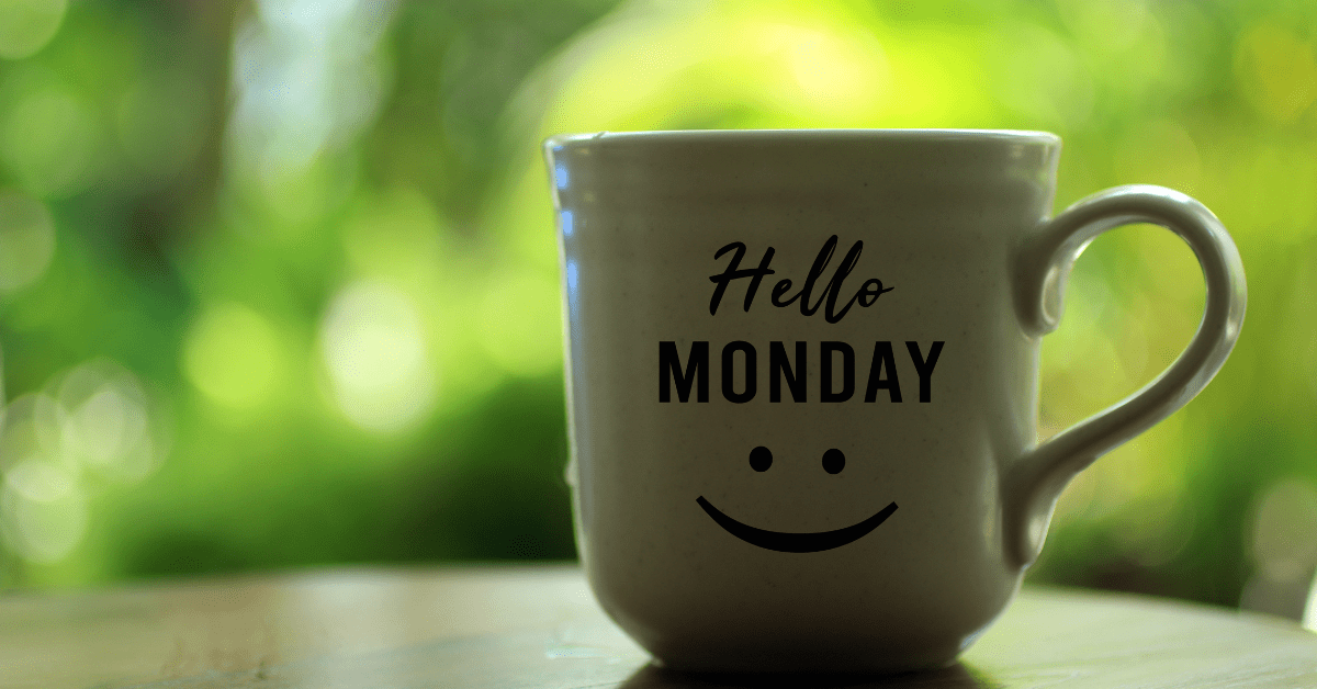 A mug on a table that has the words Hello Monday written on it with a smiley face