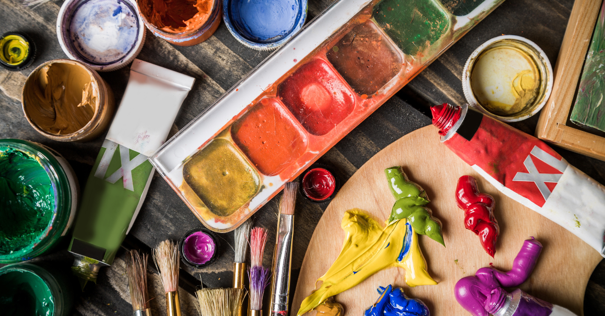 a messy desk space with various paints, pots, paint brushes and easels across it