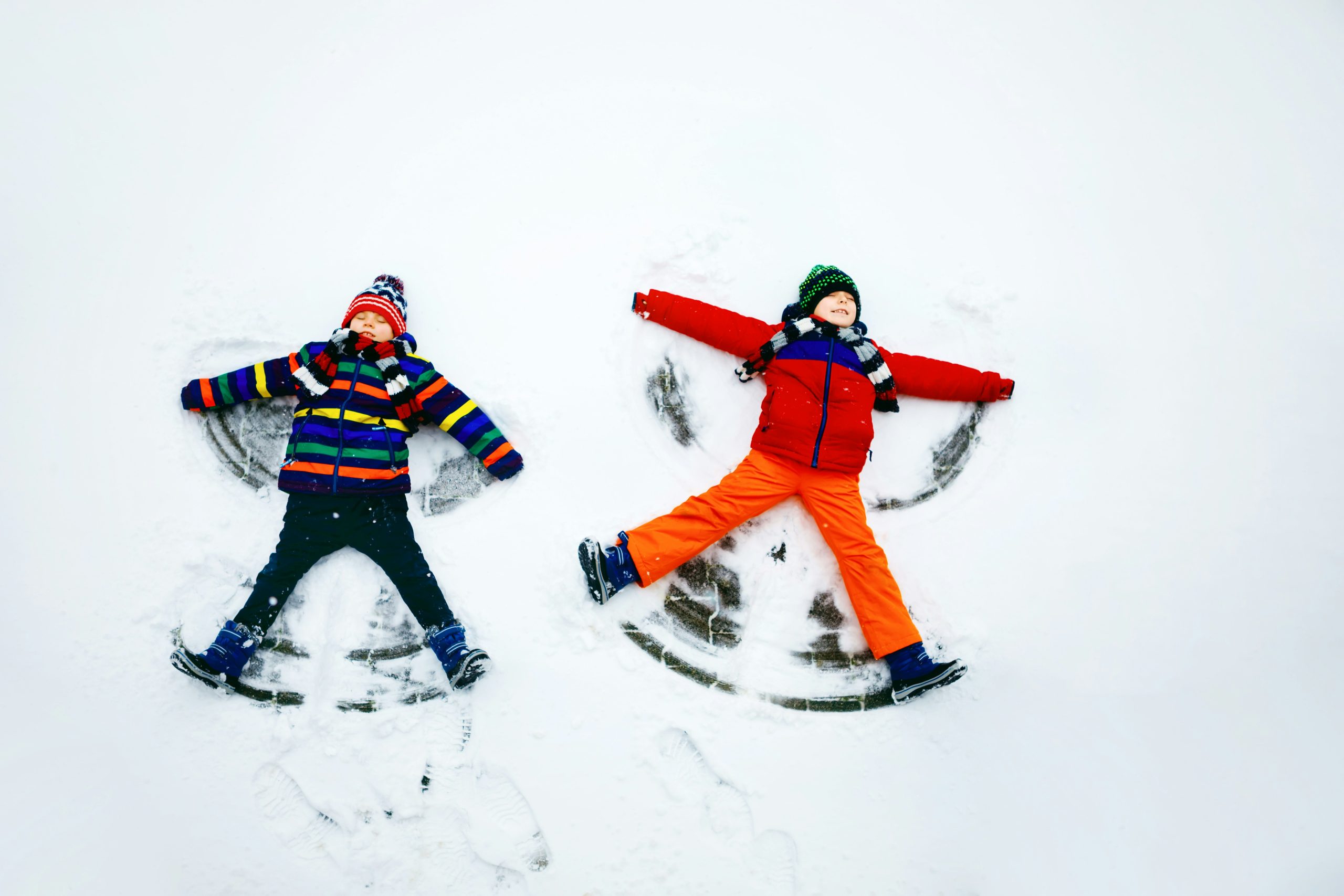 Two children in colorful winter jackets and hats lie on their backs in fresh snow, smiling as they make snow angels with their arms outstretched.