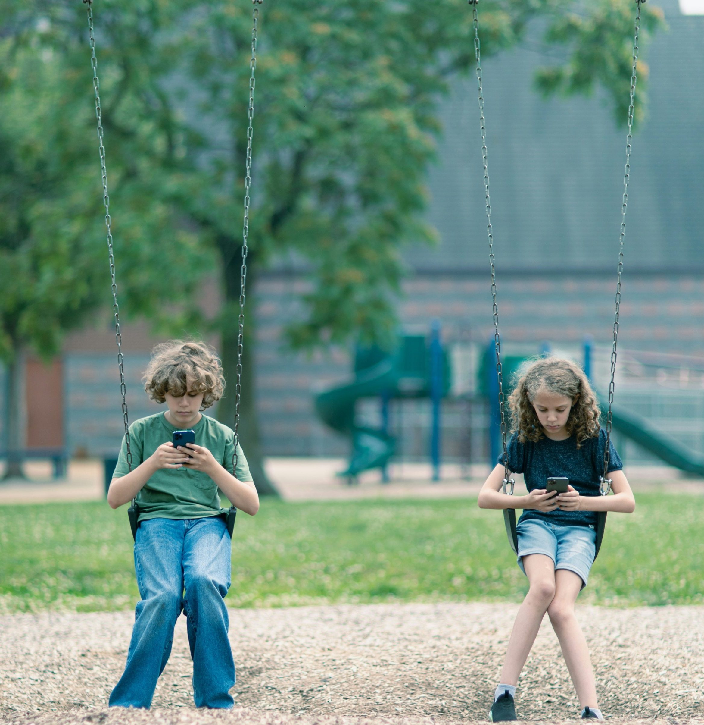 Two school-aged children sitting on swings while looking at their phones.