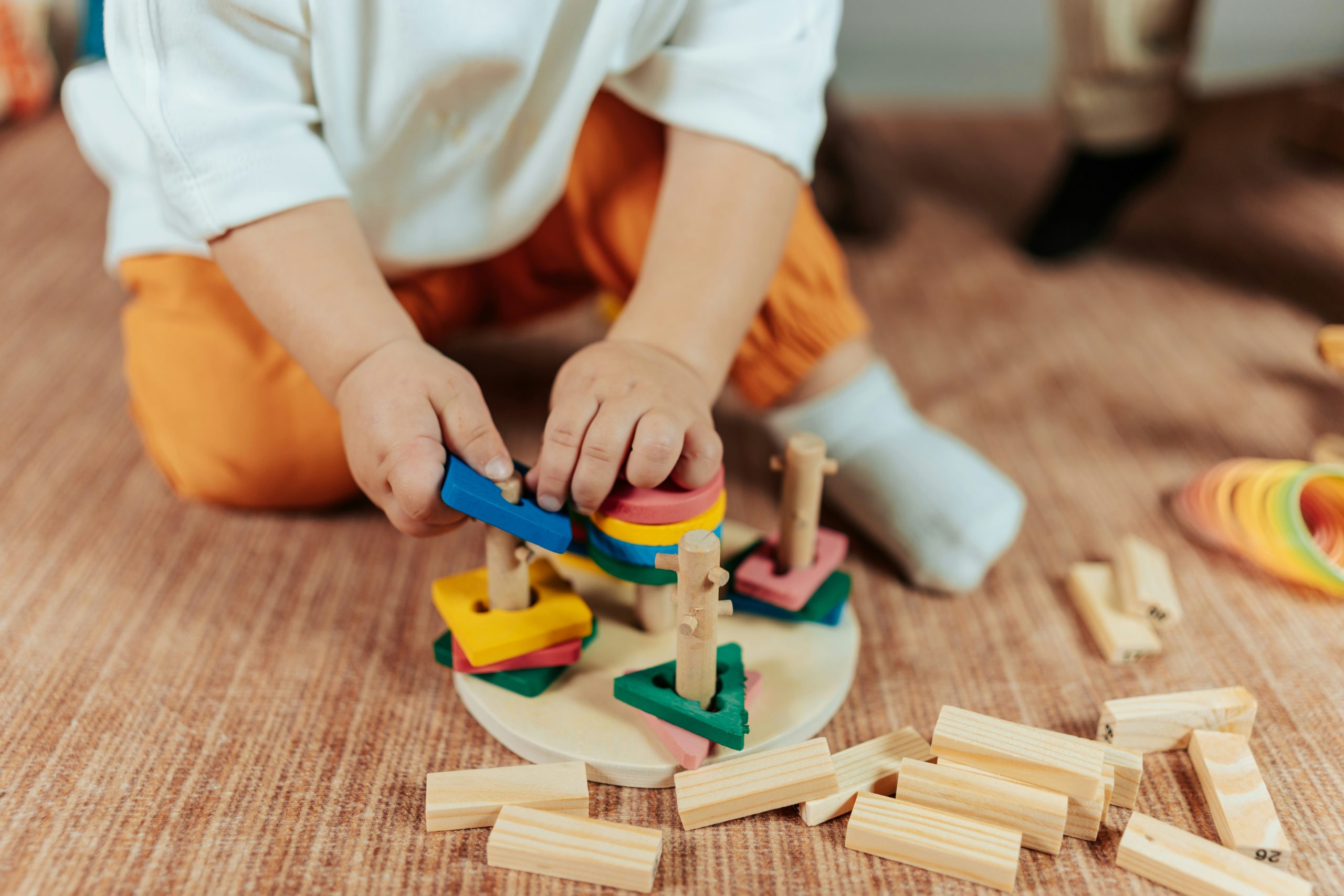 Child sitting on the floor, using a wooden shape-sorting toy, with colourful shapes and wooden blocks nearby.